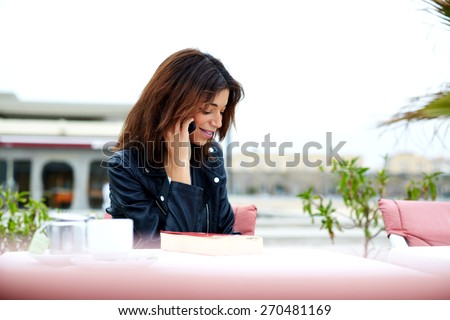Portrait of gorgeous young woman talking on mobile phone while sitting in restaurant terrace, brunette woman having cell phone conversation with someone smiling, woman relaxing on exterior terrace