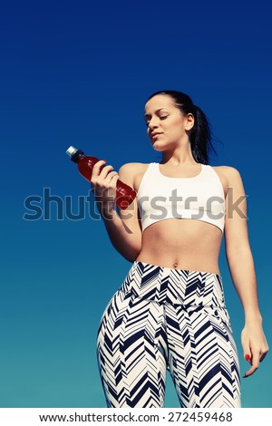 Fit woman with beautiful figure taking water break during her daily exercise routine on the beach,attractive female runner looking to the bottle of energy drink standing against sky background, filter
