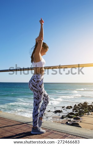 Healthy young woman stretching before her run while standing on wooden jetty with beautiful beach coastline on background,female runner warming up before a workout outdoors at sunny summer day outside