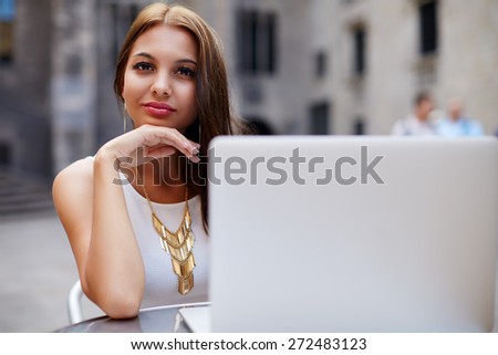 Smiling gorgeous woman sitting at street coffee shop looking to you, businesswoman sitting with open notebook next to her, female student read digital book on laptop computer sitting in sidewalk cafe