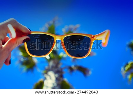 Female hand holding colorful sunglasses against palm tree and blue sunny sky, summer vacation holidays concept, first person shot, looking though glasses