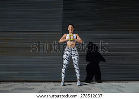 Athletic female holding weights dumbbells with her hands together, young woman in sporty clothing training bicep curls outdoors standing against background with copy space for text message by sides