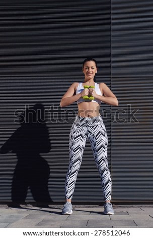 Smiling sporty woman using dumbbells to work out her arms while training outdoors against black wall with copy space for your text message, female in workout gear holding weights with hands together