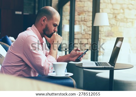 Male student work on his computer while chatting on smart phone, young business man use laptop sitting at sofa of modern coffee shop or hotel hall, freelancer working on notebook at hipster loft space