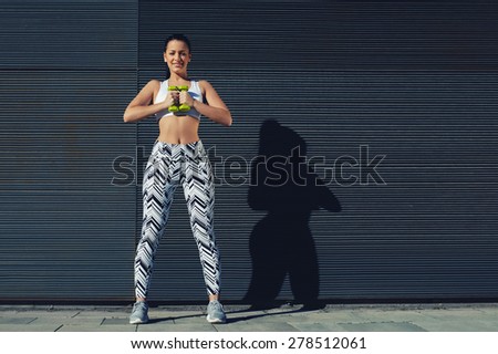 Athletic female holding weights dumbbells with her hands together, young woman in sporty clothing training bicep curls outdoors with copy space background for text message by side, filtered image