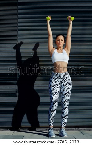 Sporty gorgeous young woman lifting weights standing with arms up against black wall background at sunset, attractive female working on her arms outdoors against copy space for your text message