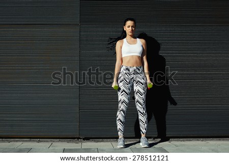 Athletic young woman holding weights with hands by sides against black wall outdoors, sexy female look to you while taking break during her training against copy space background for your text message