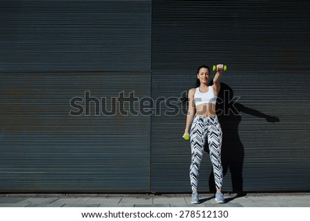 Athletic young woman lifting weights standing with arms tense against black wall outdoors, female working on her arms training bicep curls with copy space background for your text message by side