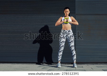 Confident sporty woman using dumbbells to work out her arms while training outdoors against black copy space wall for your text message, female in workout gear holding weights with her hands together