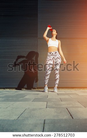 Young fit woman with beautiful figure refreshing with energy drink against black wall in the city,female runner taking break after workout standing against background with her amazing feminine shadow