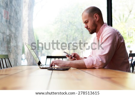Modern business man using smart phone and laptop computer in light loft interior, young hipster busy using smart phone at office desk, male student typing message on phone at wooden coffee shop table