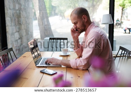 View thought vase with flowers, male freelancer working on computer and talking on smart phone while sitting at wooden table of modern coffee shop space, business man speaking on phone at office