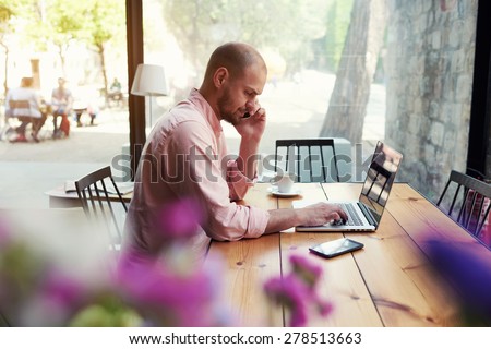 Male student work on computer touch pad while talking on smart phone, young business man use laptop sitting at wooden table of modern coffee shop, freelancer working on notebook at hipster loft space