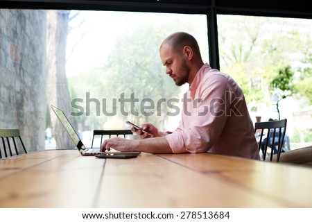Male student typing text on computer keyboard and hold smart phone, young business man use laptop sitting at wooden table of modern coffee shop, freelancer working on notebook at hipster loft space
