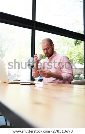Male freelancer connecting to wireless on his smart phone during coffee break, modern business man typing text message oh cell phone while working in loft studio, university student working at cafe