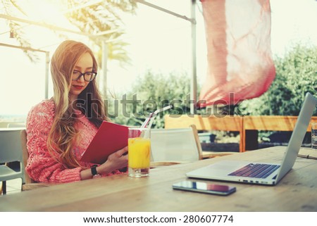 Attractive female freelancer reading notebook while sitting at wooden table front open computer in modern coffee shop, young creative woman work on laptop while having breakfast on terrace, flare sun