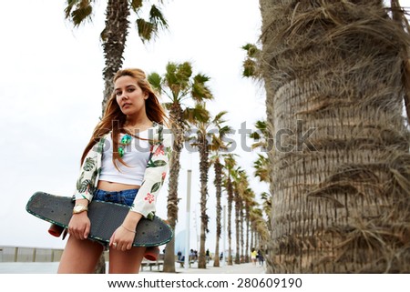 Portrait of young hipster girl holding skateboard while she standing on the lane lined with beautiful palm trees, cool trendy woman posing with her long board during recreation promenade in summer