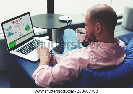Experienced businessman sitting front laptop computer with financial information as graphics and charts, young entrepreneur work on notebook drinking cafe of cup while he sitting in modern coffee shop