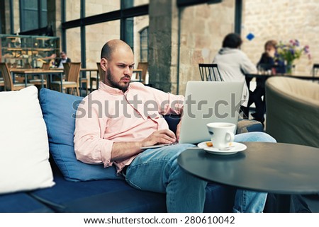 Young freelancer sitting at sofa while thoughtfully work on his notebook, successful businessman having coffee break while read some news on laptop computer with cup of cafe or tea on the table