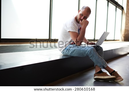 Young man looking thoughtful while working on laptop computer holding it on the knees, university student using notebook for write his coursework, freelancer sitting in modern interior thinking about