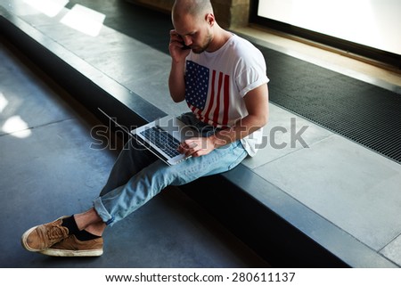 Young modern businessman connecting to wireless on laptop computer while talk on his smart phone, male freelancer working busy on notebook while having conversation on cell phone in loft studio