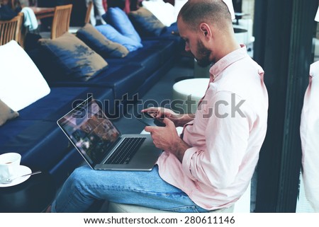 Male student work on his notebook while chatting on smart phone, young business man use laptop computer sitting in modern coffee shop or hotel hall, freelancer working on-line at hipster loft space