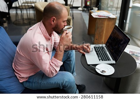 Successful freelancer drink his coffee while work on laptop computer in modern interior, young businessman connecting to wireless on notebook during breakfast in cafe,stylish hipster use modern device