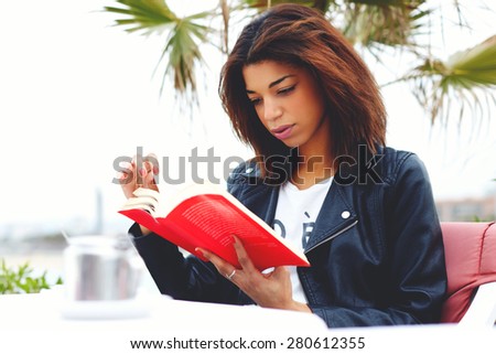 Attractive afro-american female reading fascinating book while sitting on the terrace of coffee shop, young brunette woman relaxing at weekend while read interesting book outdoors in cafe restaurant