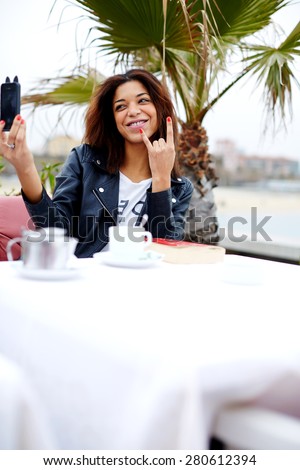 Afro american female taking fun self portrait with smart phone camera while sitting at cafe terrace near the beach, hipster young woman taking a picture of herself on her cell phone looking playful