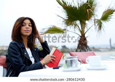 Carefree young woman enjoying a good day with book while sitting at sidewalk cafe near the beach, attractive afro american woman looking for someone in restaurant