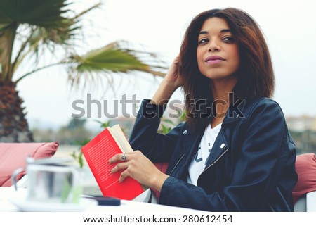 Portrait of charming afro american woman sitting at the coffee shop terrace with book or novel looking carefree and happy, female hipster enjoying her recreation time at weekend, filtered image
