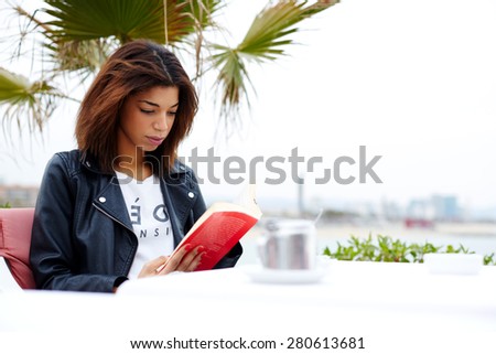 Attractive afro american woman enjoying a interesting book or novel while sitting at cafe terrace, young female hipster fascinating read book in open air coffee shop during her recreation time