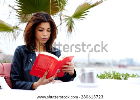 Attractive afro american woman enjoying a interesting book or novel while sitting at cafe terrace, young female hipster fascinating read book in open air coffee shop during her recreation time