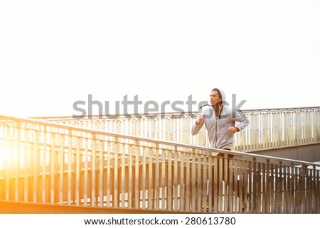 Sporty young man working out at early morning while run up the bridge with orange sunrise on background, male jogger exercising while listening to music with headphones, runner working out outdoors
