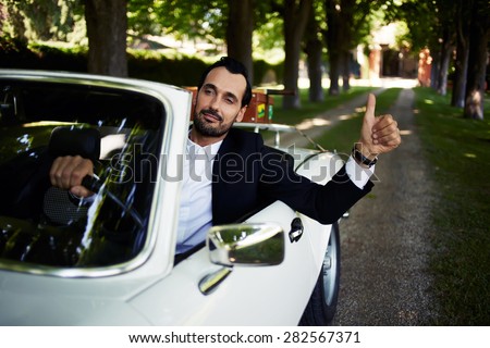 Successful and wealthy businessman sitting behind steering wheel of luxury cabriolet car on countryside road, confident handsome man sits in his new convertible car doing super gesture with the finger