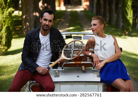Happily married couple enjoying a beautiful day outdoors while leaning on their convertible antique car, handsome wealthy man and his gorgeous female posing outdoors, people spending time together