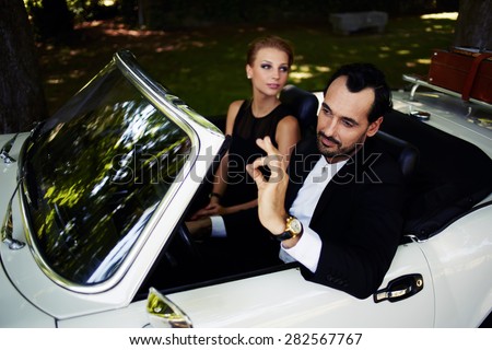 Richly dressed wealthy and famous couple sitting inside cabriolet luxury car, successful man sitting behind convertible steering wheel with its ok hand gesture, happy man and woman enjoying the life