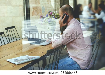 View though window, young businessman using laptop computer and smart phone while working busy, male student or freelancer working on notebook at wooden table of modern coffee shop, glassy reflection
