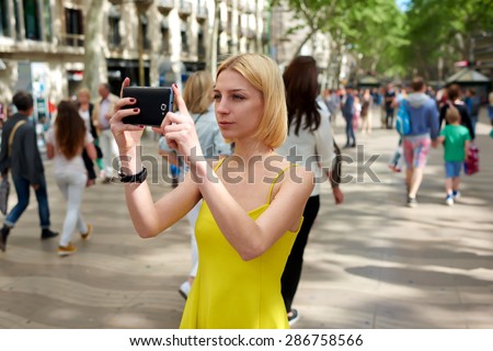 Pretty young woman photographing urban view with mobile phone camera during summer journey, gorgeous female tourist taking picture with her smart phone while standing outdoors in urban setting street