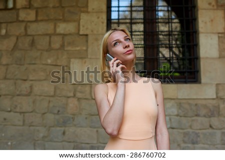 Gorgeous young woman talking on her smart phone while standing in city street with copy space wall,female hipster having cell phone conversation looking up thoughtfully,businesswoman at telephone call