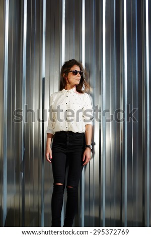 Trendy young female hipster dressed in a classy modern clothes standing on a shiny metal background with copy space for your text message or content and look to the aside on a sunny summer day