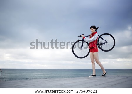 Full length portrait of flamboyant stylish girl dressed in a red sport jacket and shorts holding her light weight fixed gear bicycle while standing on concrete pier seashore with copy space cloudy sky