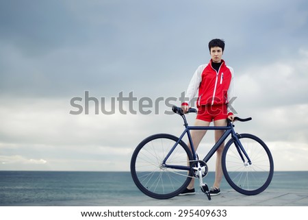 Full length portrait of stylish hipster girl standing with her sport fixed gear bike on the beach, young woman standing on the pier before ride along the seaside on her bicycle,promenade at cloudy day