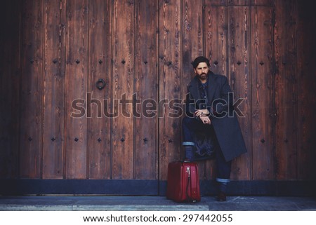 Portrait of stylish bearded confident man with vintage suitcase standing on a wooden wall background with copy space for your text message or content,male model with confident look posing outdoors