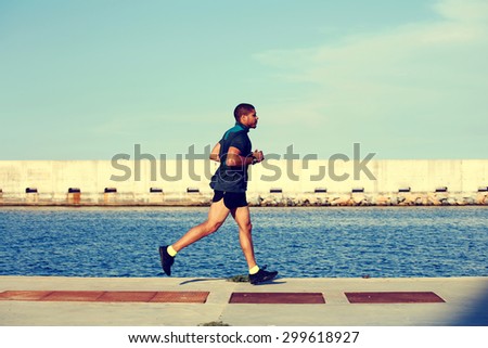 Young muscular runner at evening jog enjoying sunny afternoon outdoors, afro american man working out in marina port against sea background with copy space area for your text message or advertising