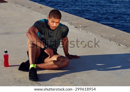 Male runner sitting on the road while taking break after workout outdoors, sportsman resting after fit training at sunny afternoon in summer,tired sweaty male jogger having rest after exercise and run
