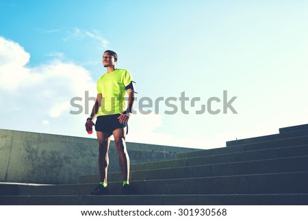 Portrait of a runner man with armband on the arm rest after jogging while standing on ladder against the sky background with copy space area for your text message information,sportsman taking a break