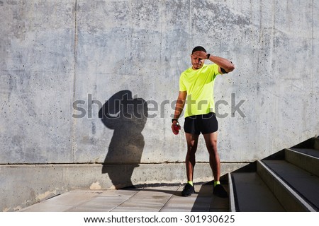 Full length portrait of sweaty man jogger rubbing his forehead while standing against cement wall background with copy space area for your text message or content, athlete resting after fit exercise