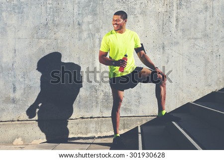 Smiling male runner in bright t-shirt holding bottle of energy drink while standing against cement wall background with copy space area for your advertising, happy afro american athlete having a rest