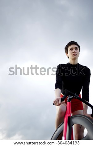 Low view portrait of young female cyclist standing with modern fixed gear bike on gloomy sky background while resting after long ride outdoors, cyclist with copy space for text message or advertising